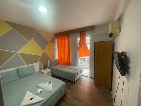 A hotel room with two beds, a small table, a TV, and a window with orange curtains.