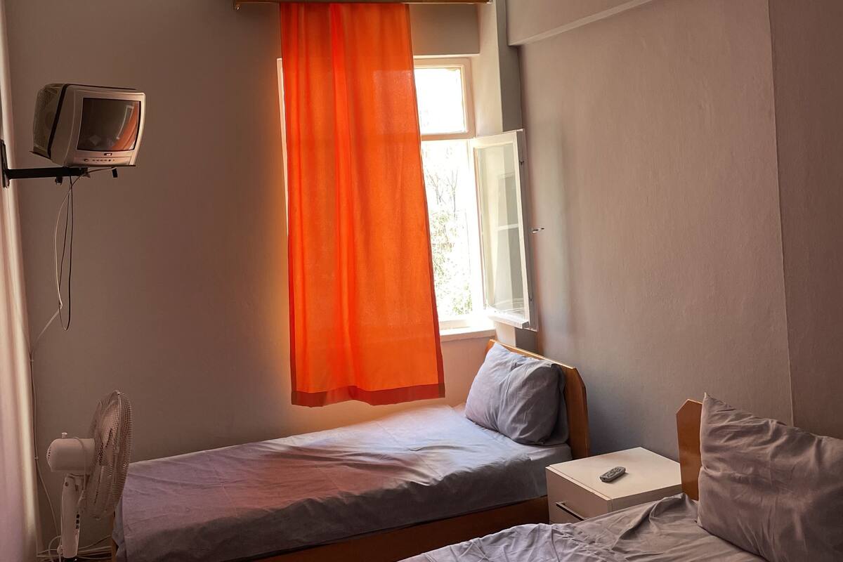 A room with two beds, a ceiling fan, a TV, and a window with orange curtains.