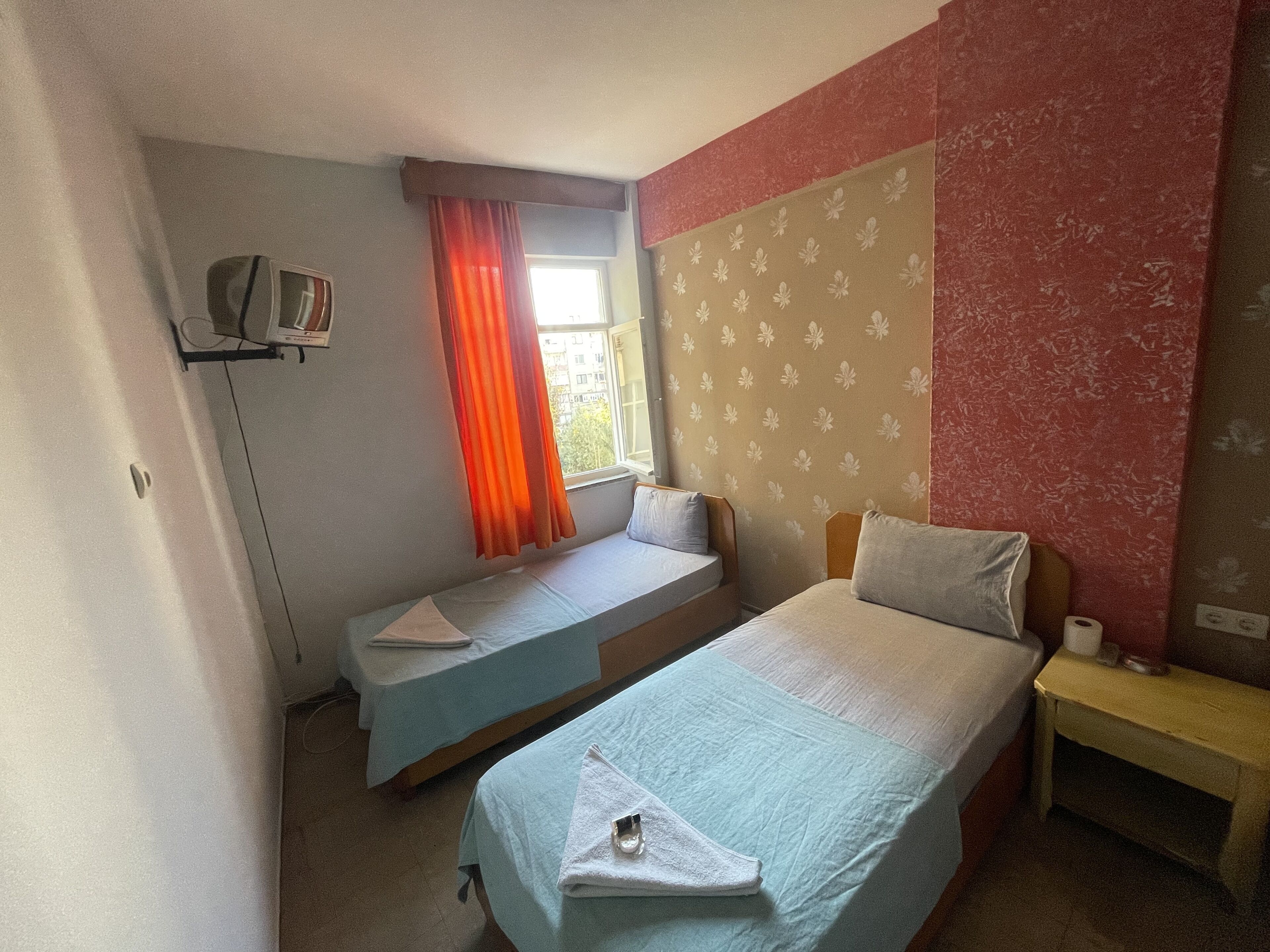 Quarto de hotel pequeno e individual, com duas camas de solteiro, uma janela com cortinas laranja, uma televisão sobre um suporte e uma parede com padrão floral.