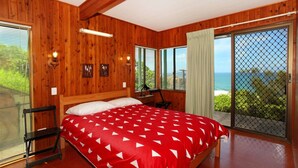 Een slaapkamer met houten inrichting, een bed met een rood-wit gebeddeerde spreidek en een bureau met stoel, plus een groot raam met uitzicht op zee.