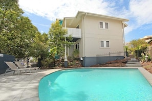 A swimming pool with lounge chairs, a two-story building with a balcony, and a fenced garden area.