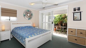 A bedroom with a bed, bedside tables, a ceiling fan, a decorative wall piece, and a view of a patio with plants and a bicycle. - Unit 3 'On the Park' at Coolum Beach (Coolum Beach)