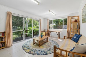 Une pièce dotée d’une grande porte-fenêtre coulissante donnant sur un jardin, d’un canapé en bois avec des coussins, d’une petite table en bois et d’une bibliothèque.