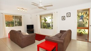 Ein Wohnzimmer mit zwei Sofas, einem roten Tisch, einem Fernseher und großen Fenstern mit Vorhängen.