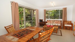 Ein geräumiges Wohnzimmer mit einem Holzespeisetisch, einer Couch und zwei Sesseln. Große Fenster bieten Blick auf Grünflächen.