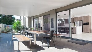 Une cuisine moderne dotée d’un four et d’un micro-ondes encastrés, un coin repas avec une table et des chaises en bois, et de grandes portes-fenêtres coulissantes donnant sur une terrasse aménagée avec du mobilier d’extérieur.