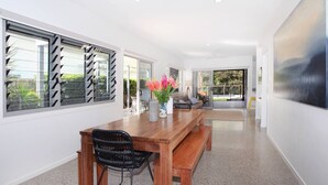 Un comedor moderno con una mesa y sillas de madera, un jarrón con flores y ventanas amplias con persianas negras.