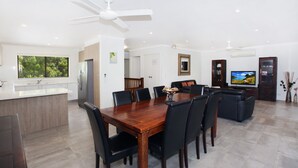 A modern kitchen with a central island, dining area with a wooden table and chairs, a living room with a sofa and TV, and a ceiling fan. - 8 Springfield Avenue (Coolum Beach)