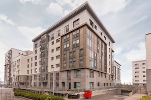 A modern multi-story building with a corner entrance, large windows, and a parking area with a red dumpster.