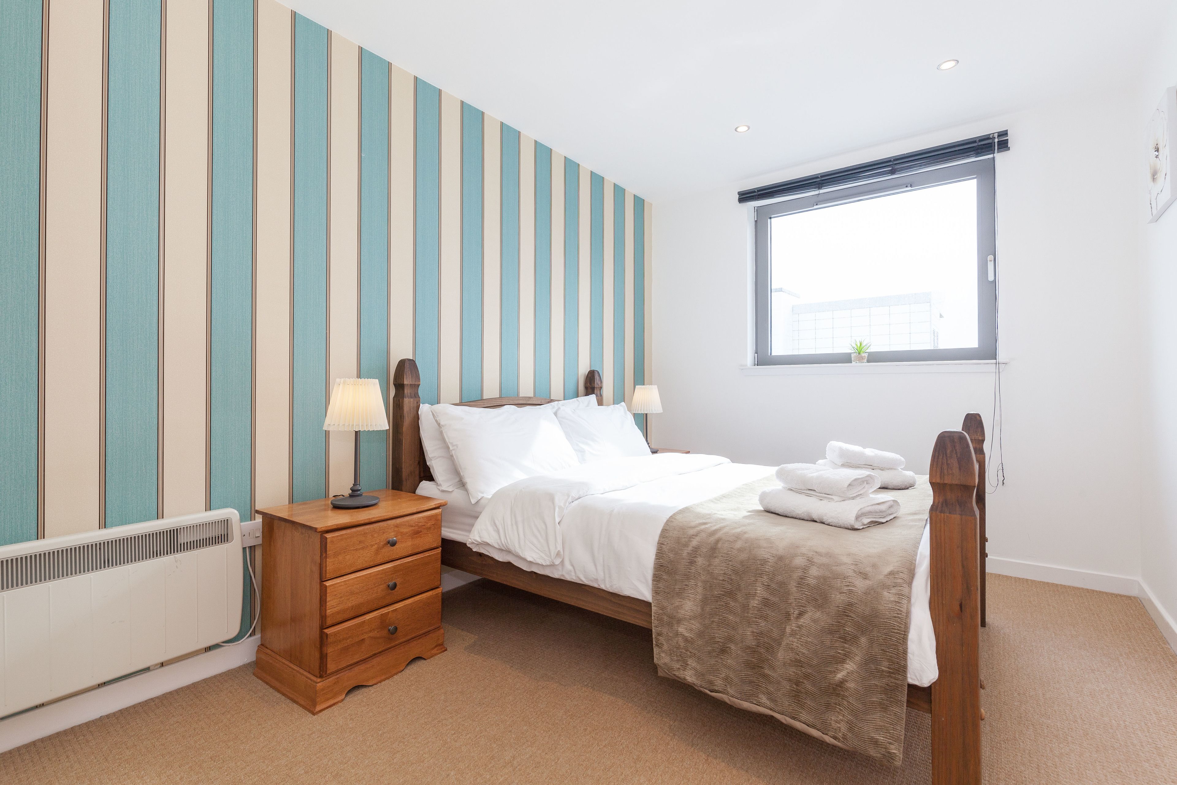 A bedroom with a bed, wooden nightstand, bedside lamps, a radiator, and a window with blinds.