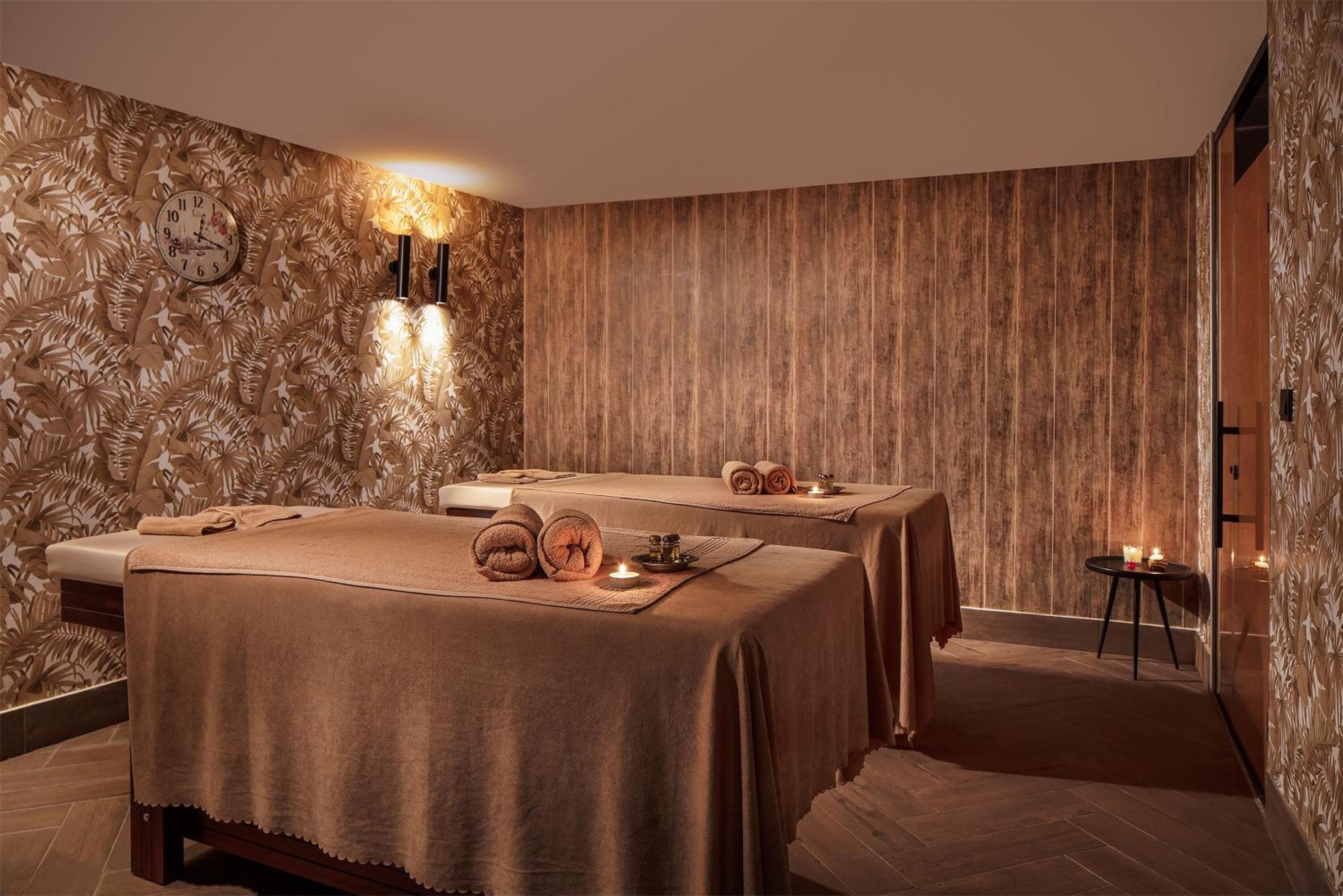 Une salle de spa équipée de deux tables de massage, d’une horloge murale et d’un mur décoratif orné de motifs de feuilles.