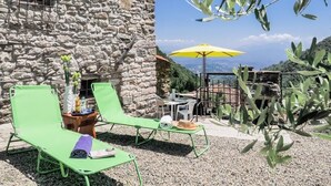 Een stenen gebouw met een terras, groene lounge stoelen, een gele parasol en een tafel met stoelen.