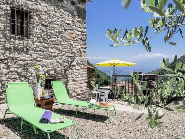 Un edificio de piedra con una terraza, sillones verdes, una sombrilla amarilla y una mesa con sillas.