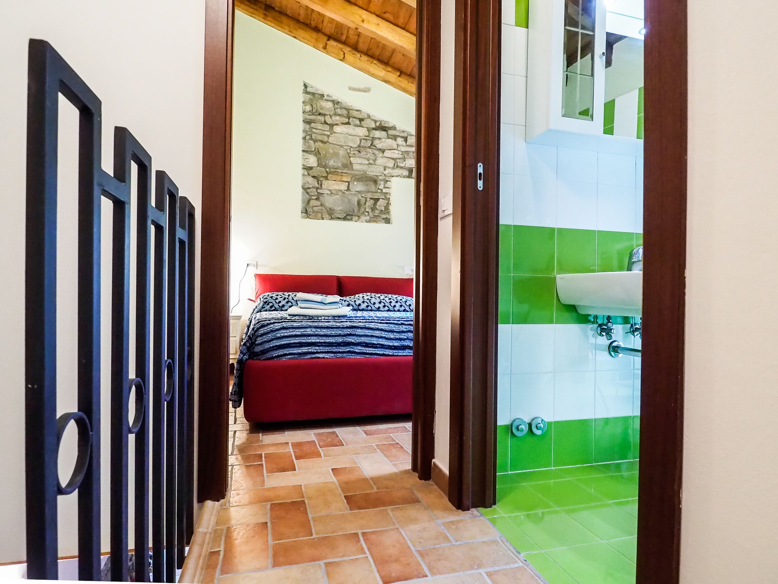 A bedroom with a red bed, a stone wall, and a bathroom with green tiles.