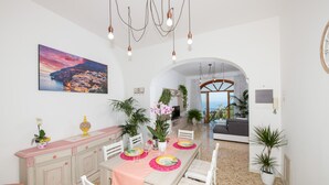 A dining area with a table set for four, surrounded by white chairs. There is a large painting on the wall, a plant on the table, and a view of the living area through an archway. - Positano Villa Genny and Emily breakfast & experience (Positano,Montepertuso)