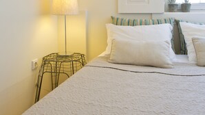 A bedroom with a bed, a lamp, two hats on the wall, and a window with plants. - Family Matters | Historic Center (Porto)