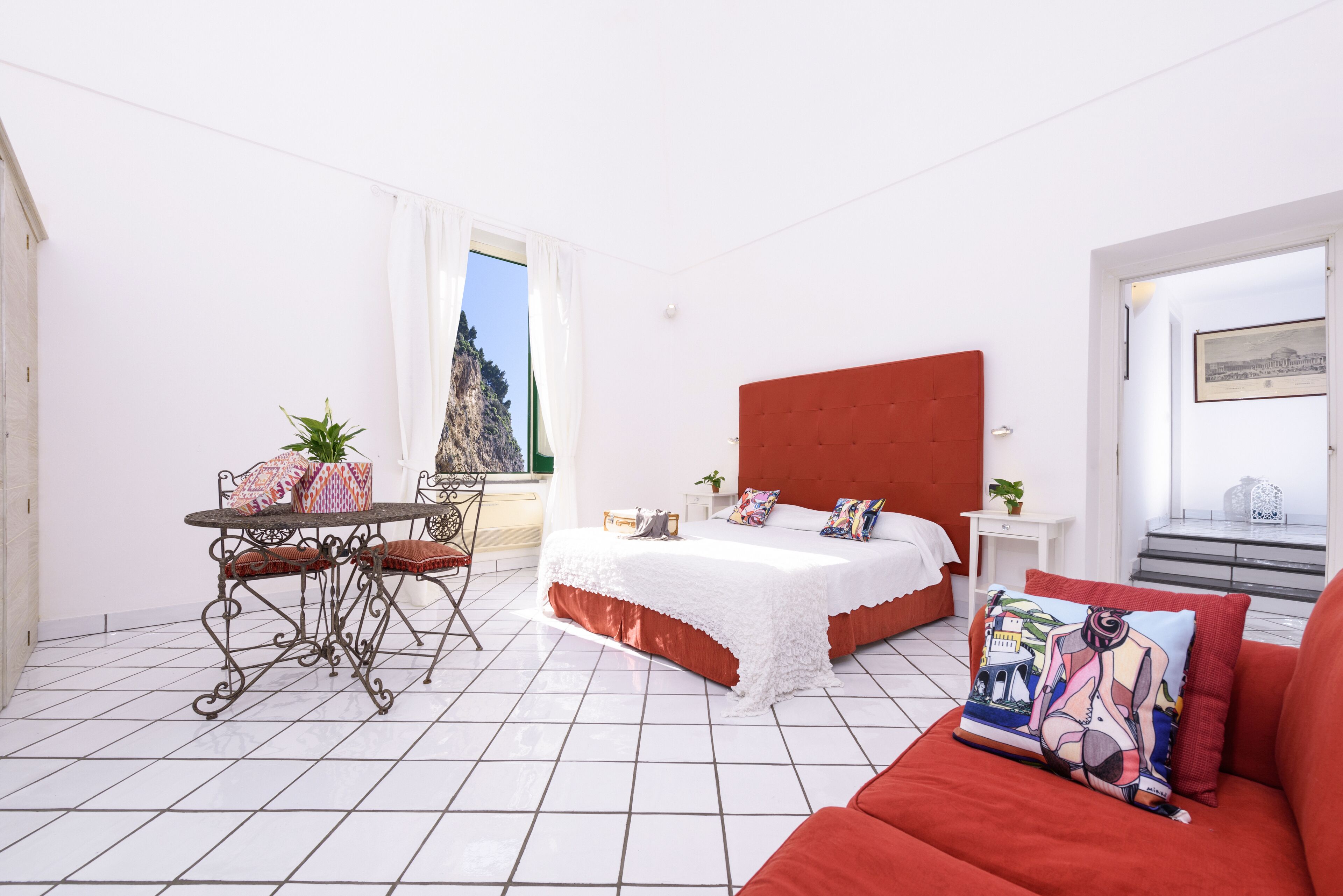 A bedroom with a bed, a red sofa, a table with chairs, a window with a view, and a white wall.