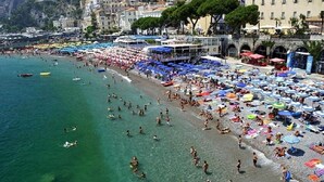 Una playa concurrida, gente nadando y tomando el sol, rodeada de sombrillas coloridas y cafés frente al mar.