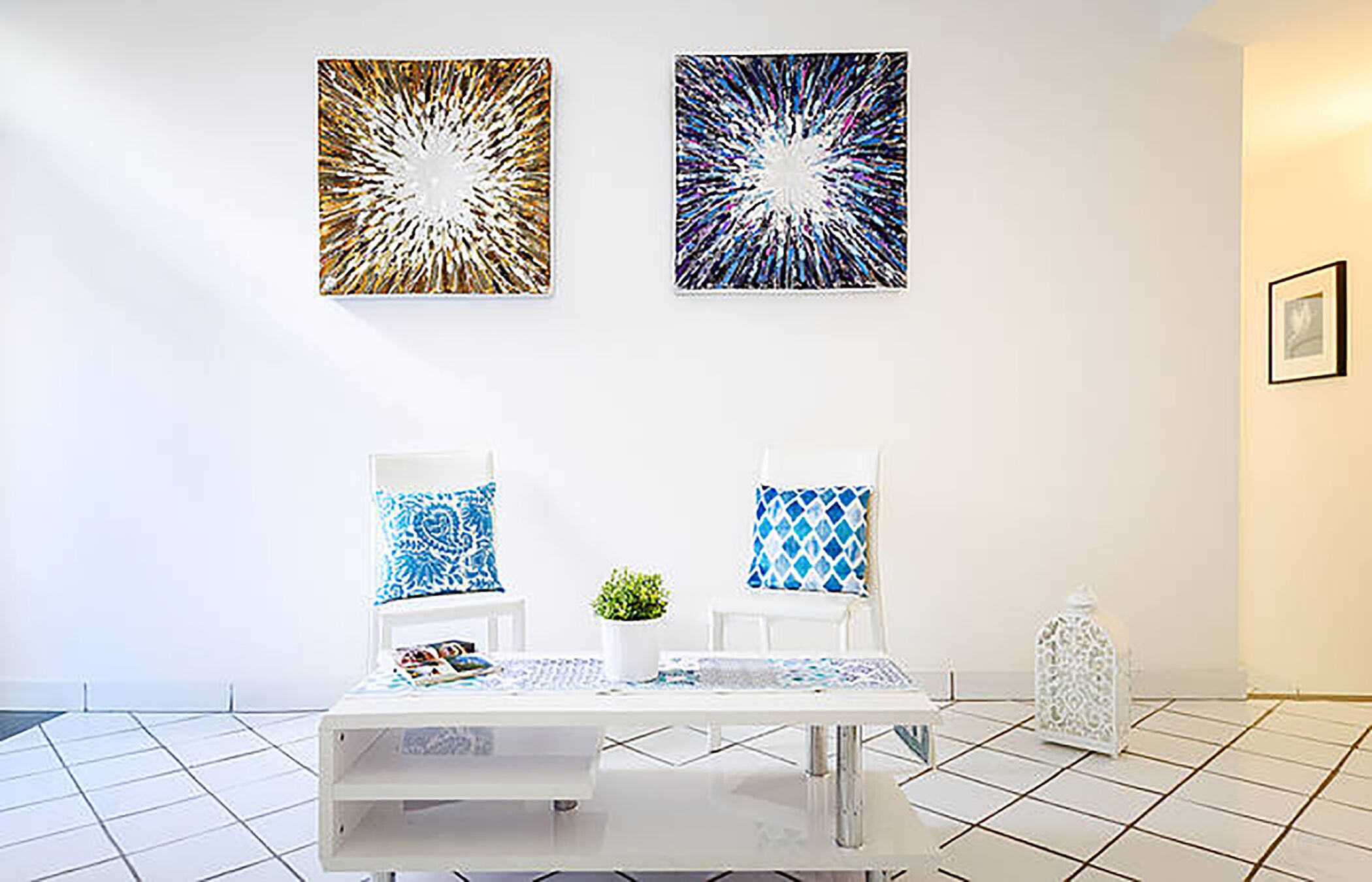 A modern room with a white table, two chairs with patterned cushions, and two abstract wall art pieces.
