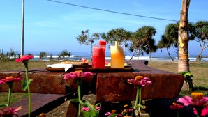 Una mesa de madera con comida y bebidas, con vista a una playa con palmeras y el océano.