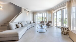 A spacious living room with a grey sectional sofa, a round coffee table, and two armchairs. A staircase is visible on the left, and large windows provide natural light and a view of the outdoors.