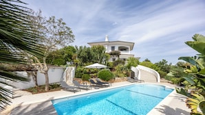 A swimming pool with lounge chairs, a white building with a balcony, and surrounding greenery.