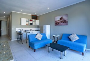 Ein modernes Wohnzimmer mit einem blauen Sofagarnitur, einem Couchtisch und einer Küchenzeile mit weißen Schränken und einem Kühlschrank.