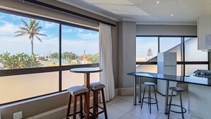 A modern kitchen with a large window, a dining area with stools, and a view of palm trees and buildings. - Ballito Bay holiday Apartment (Ballito)