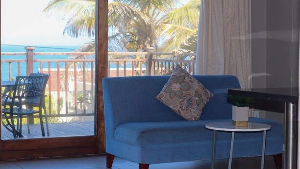 A room with a blue sofa, a small round table, and a view of the ocean and palm trees through a large window. - Ballito Bay holiday Apartment (Ballito)
