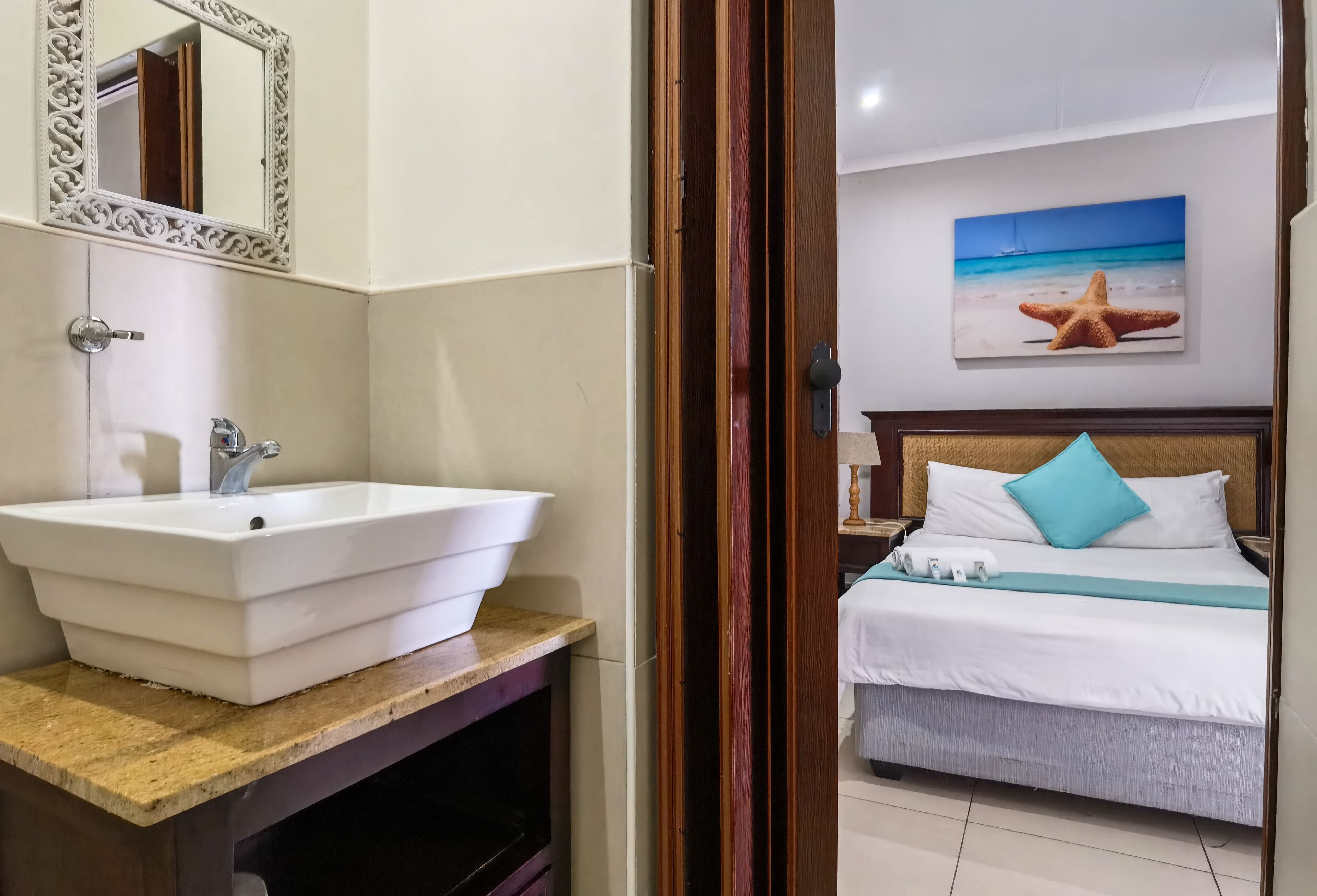 A bathroom with a white sink and mirror, a wooden vanity, and a bed with white bedding and a blue pillow.