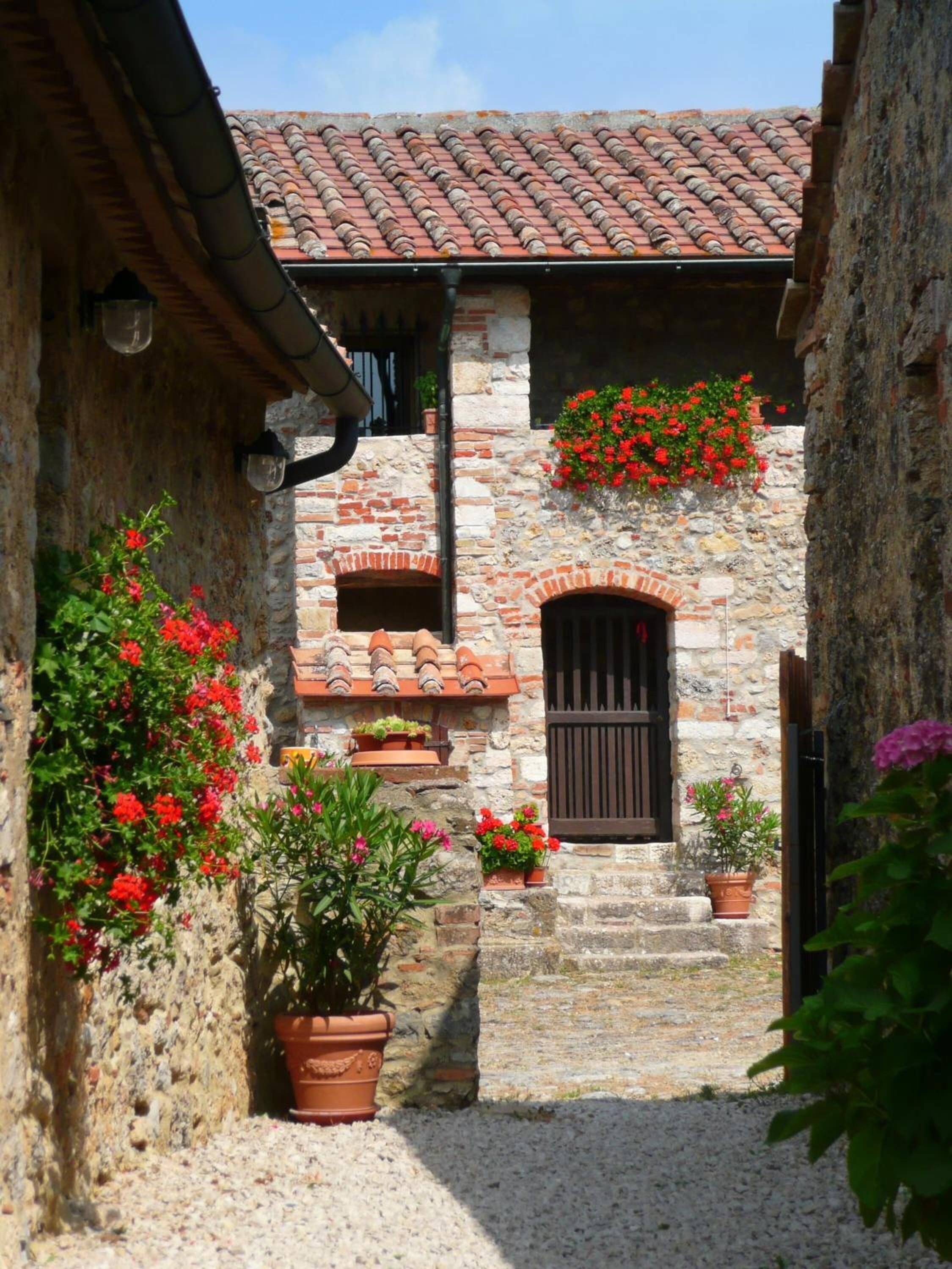Un stretto sentiero in pietra conduce a un edificio rustico in pietra, con tetto in tegole, piante in vaso e una porta in legno.