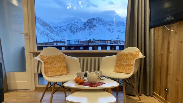 Una habitació acollidora amb dues cadires blanques i una taula petita, una finestra amb vista a muntanyes cobertes de neu, i un televisor penjat a la paret.