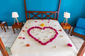 Habitación de hotel con cama, dos mesitas de noche con lámparas, una silla azul y un arreglo floral en forma de corazón sobre la cama.