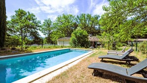Una piscina rectangular con superficie de agua azul cristalina, rodeada por una zona cubierta de césped. Hay dos sillas de descanso con cojines oscuros y estructura metálica.