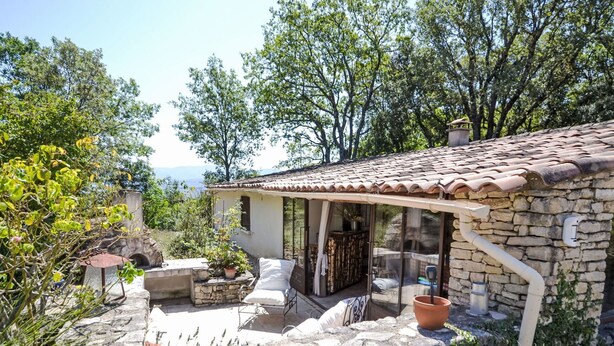 Una casa de piedra con techo de tejas, un porche cubierto y vistas de los árboles y montañas circundantes.