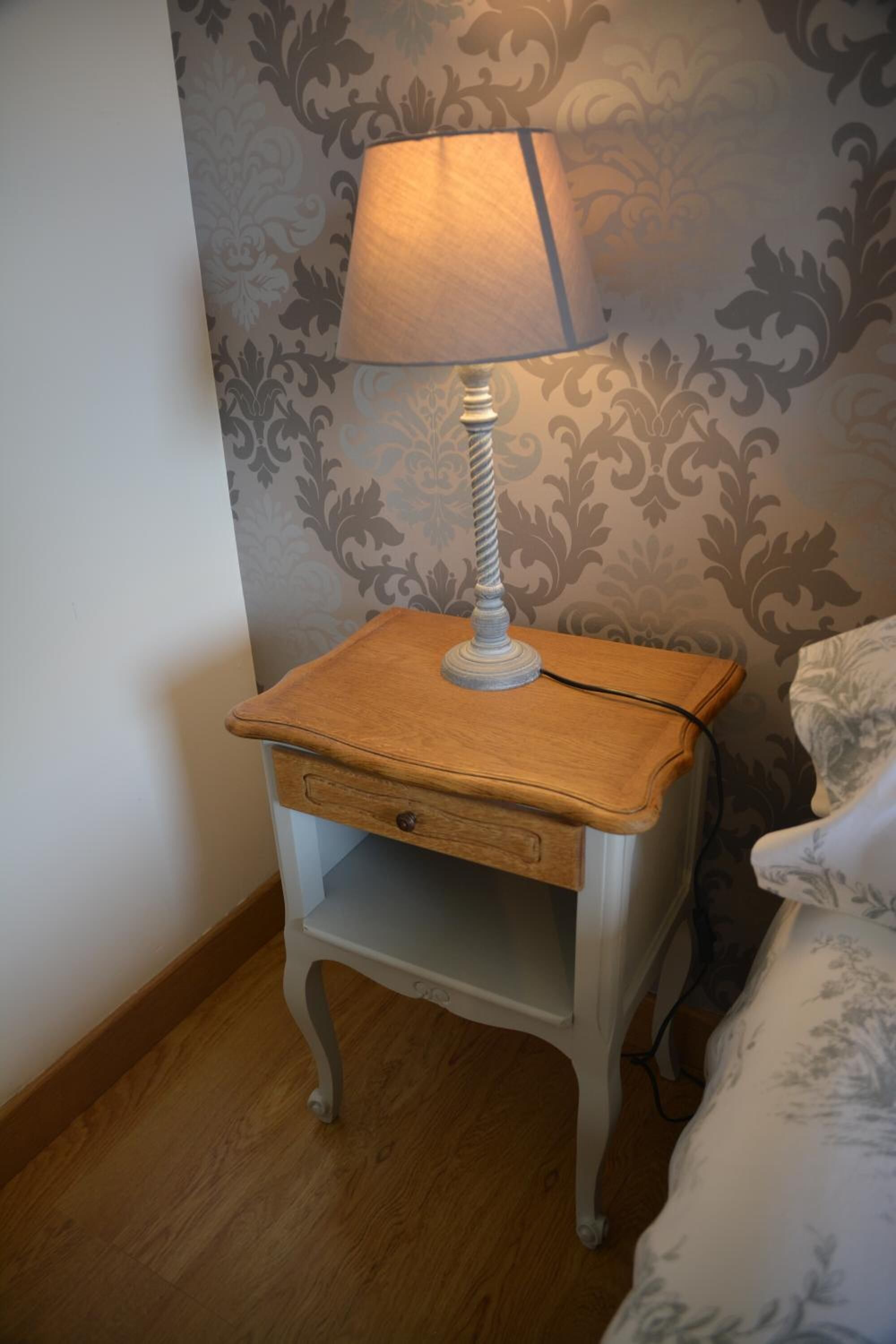 A small bedside table with a lamp, a drawer, and a decorative wall with a damask pattern.