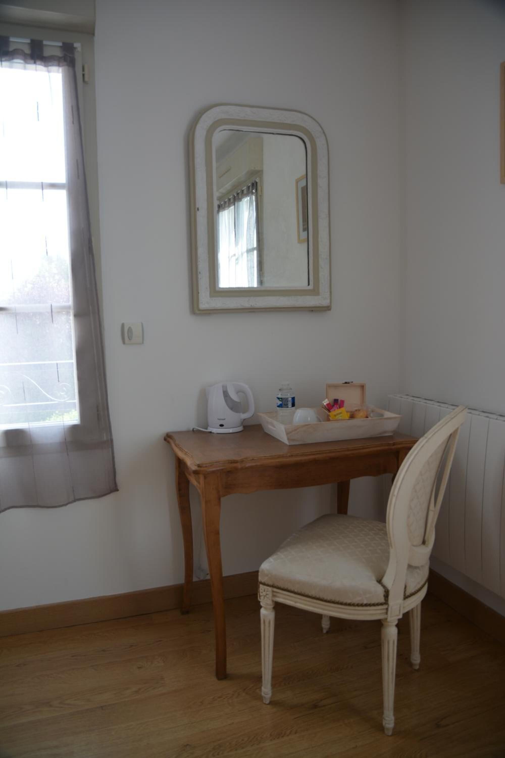 A small room with a wooden desk, a white chair, a mirror, and a window with curtains.
