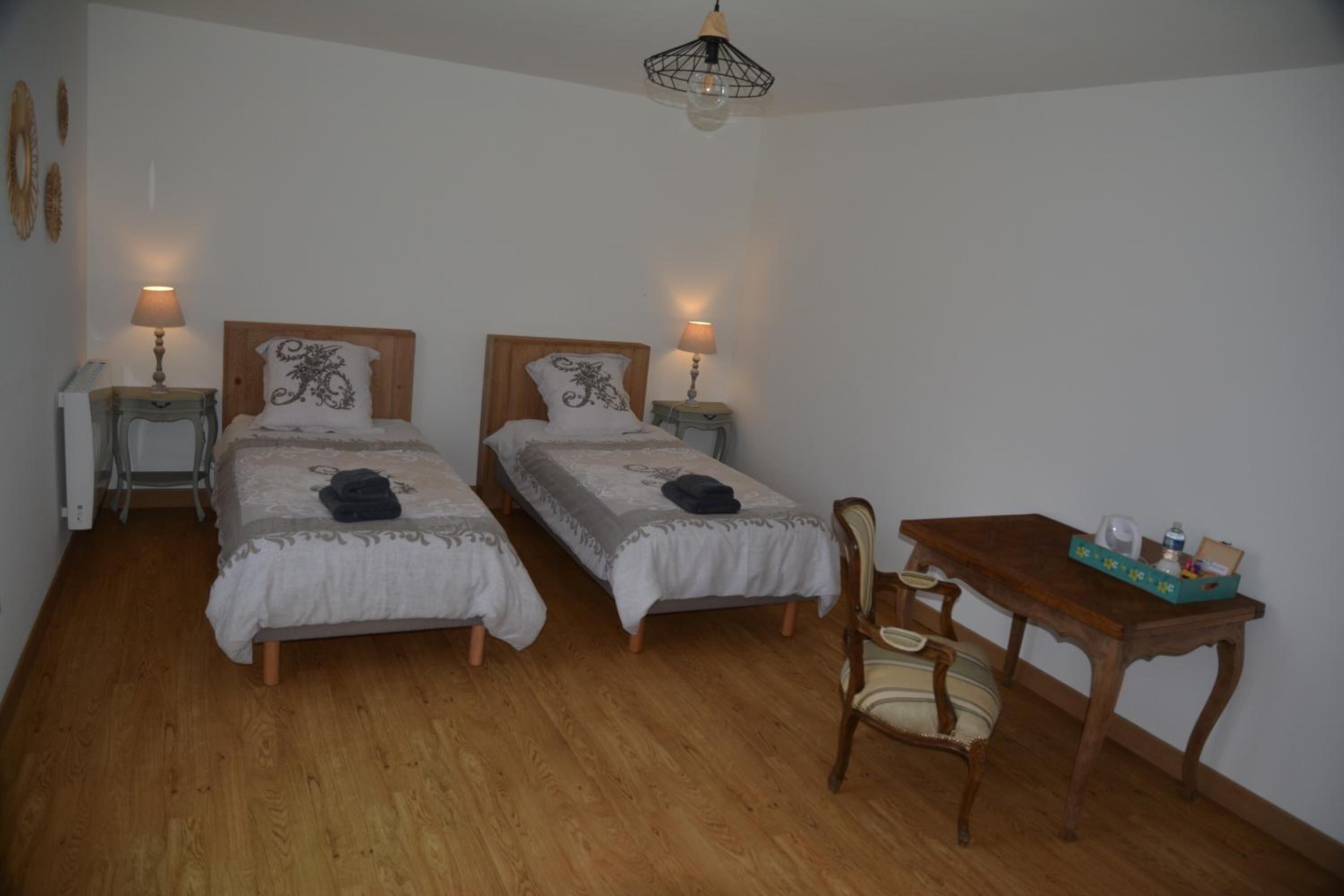 A room with two single beds, wooden nightstands with lamps, a wooden table with a chair, and a hanging light fixture.