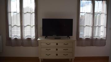 A room with a television on a dresser, two windows with curtains, and a radiator on the left.