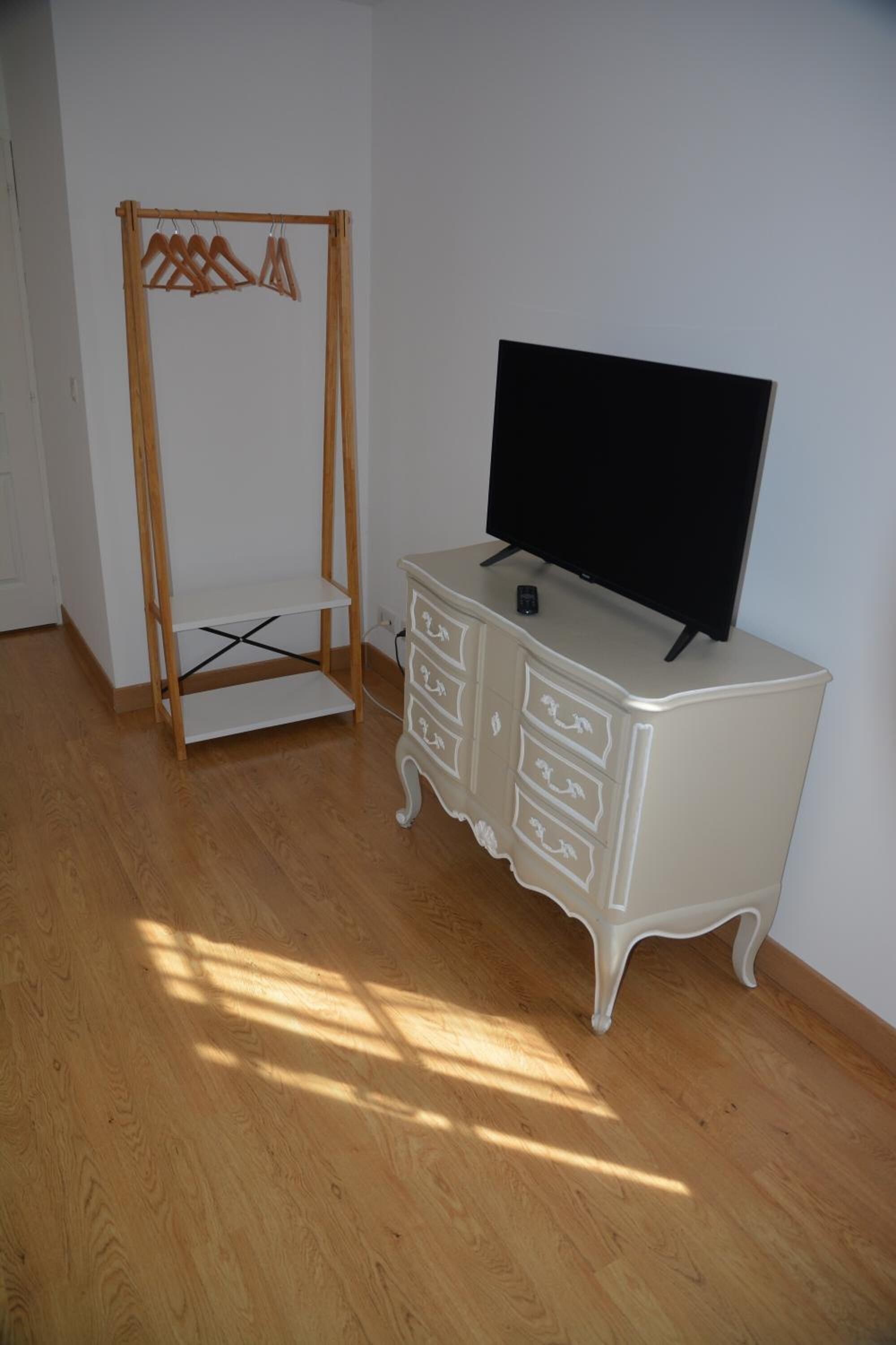 Quarto com piso de madeira, cômoda branca com gavetas, televisão por cima e um cabideiro de madeira com cabides.