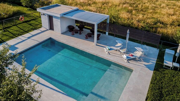 A modern outdoor pool area with a rectangular pool, a covered seating area with chairs, and a lounge area with sun loungers.
