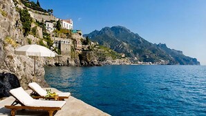 Eine Küstenansicht mit einer Betonplattform, zwei Liegestühlen mit weißen Kissen, einem weißen Sonnenschirm und einer felsigen Klippe mit Gebäuden.