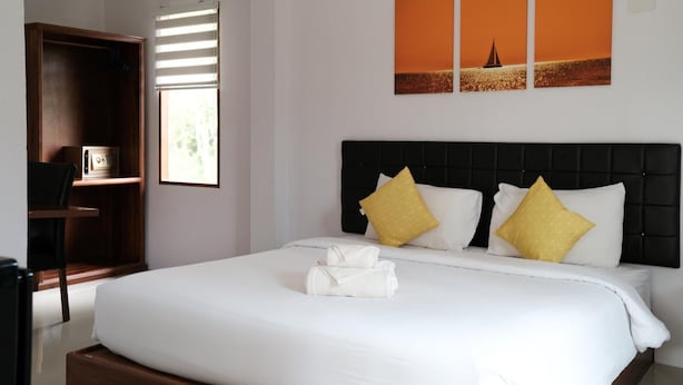 Una cama bien tendida con sábanas blancas y dos almohadas amarillas, un armario de madera y una ventana con persianas.