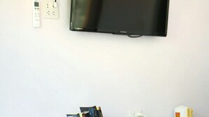 Un espacio habitable compacto con un televisor de pantalla plana montado en la pared, un refrigerador pequeño, una mesita de madera con libros y una tetera, y un armario blanco con cajones.