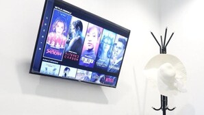 Televisor de pared mostrando contenido original de Netflix, un mueble de madera con aperitivos, una tetera y una lámpara de pie.