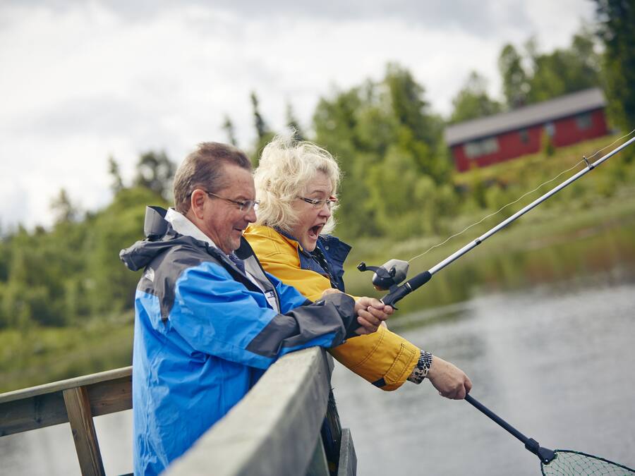 Deux personnes sont en train de pêcher sur un bateau, avec un lac et un bâtiment rouge en arrière-plan.