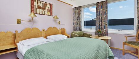A hotel room with a bed, a chair, a small table, a window with curtains, and a wall-mounted lamp.