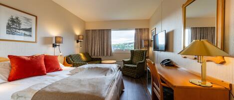 A hotel room with a bed, two chairs, a desk, a lamp, a TV, and a large window with curtains.