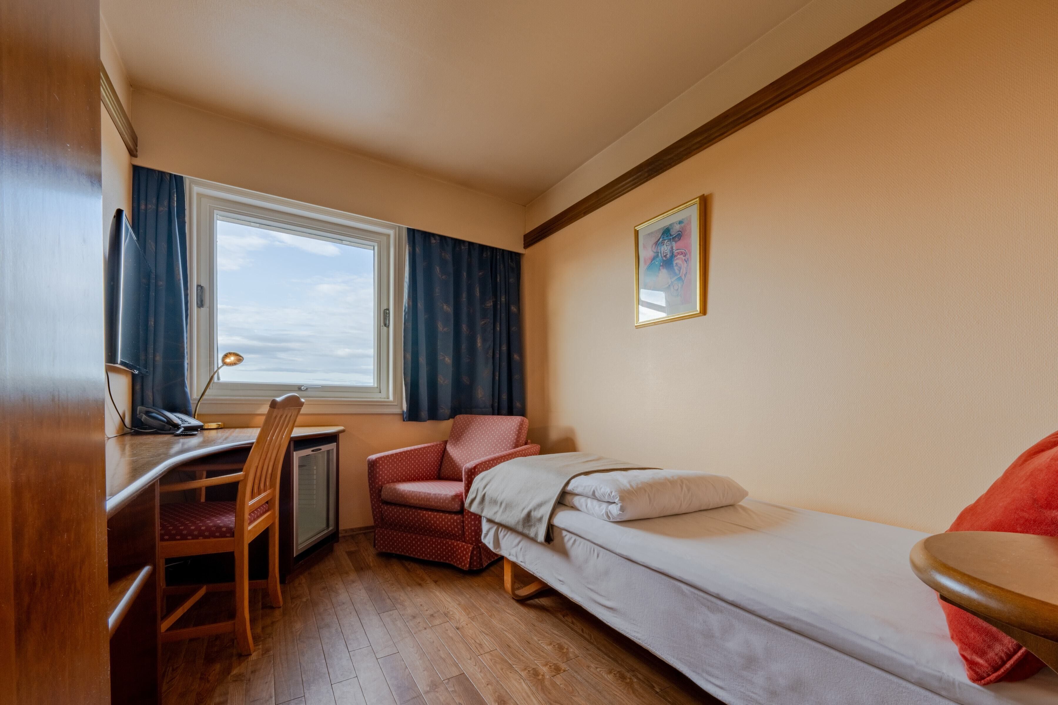 A compact hotel room with a single bed, a desk with a chair, a small table, and a window with curtains.