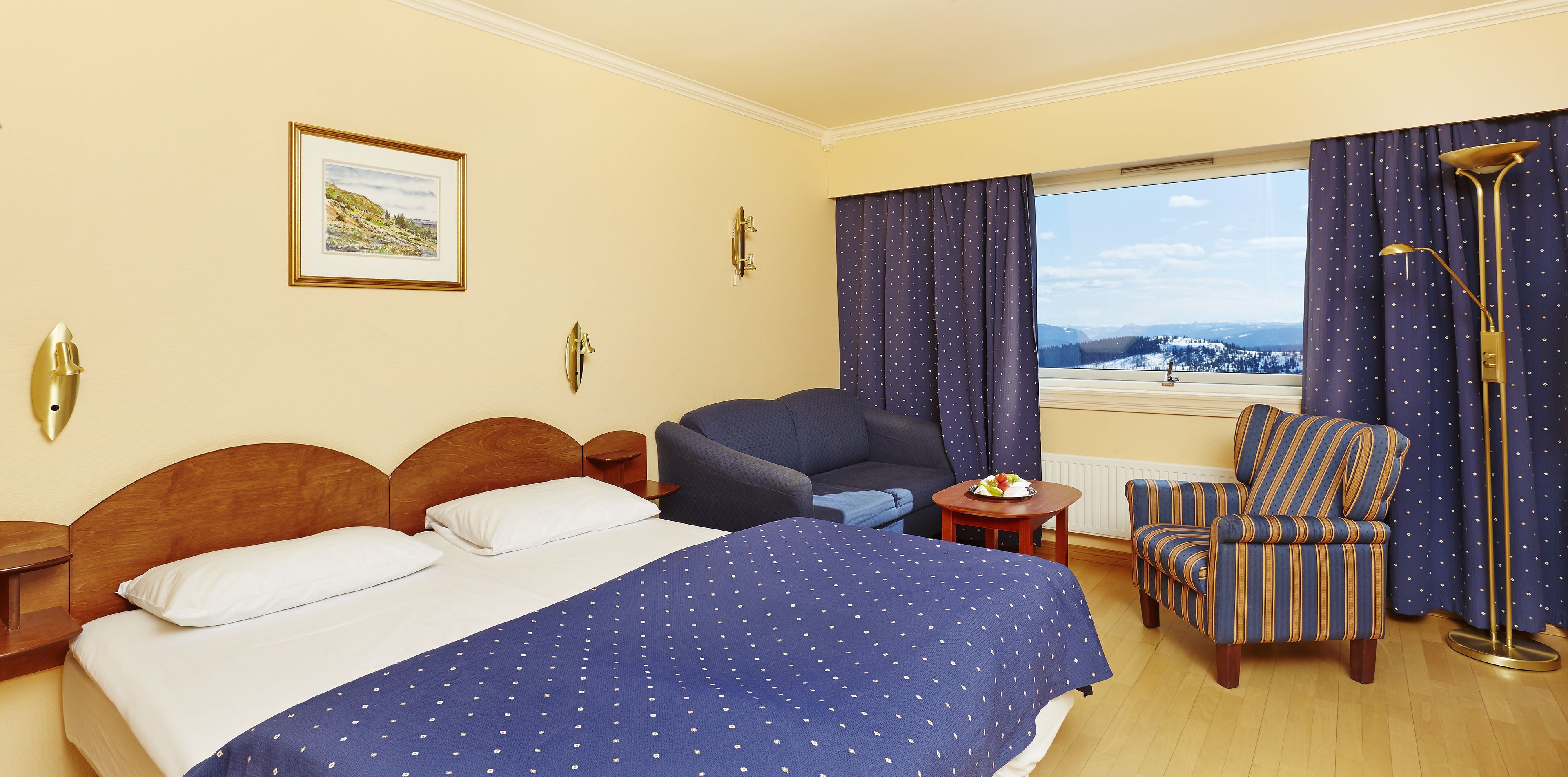 A hotel room with a bed, a chair, a small table with a plate of fruit, and a view of a snowy landscape through the window.